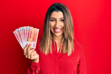 Beautiful brunette woman holding 100 norwegian krone banknotes looking positive and happy standing and smiling with a confident smile showing teeth