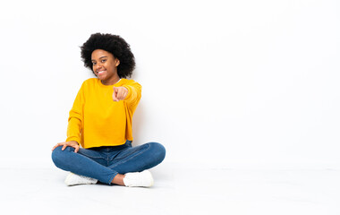 Young African American woman sitting on the floor points finger at you with a confident expression