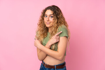 Young blonde woman with curly hair isolated on pink background pointing to the laterals having doubts