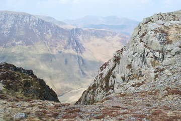 A rocky chasm; cliffs and crags in a steep valley descending dramatically from a mountain top to the valley below. Other mountains in the distance; a stream or river at the bottom of the valley