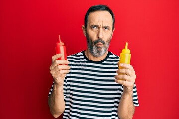 Middle age hispanic man holding ketchup and mustard bottle making fish face with mouth and squinting eyes, crazy and comical.