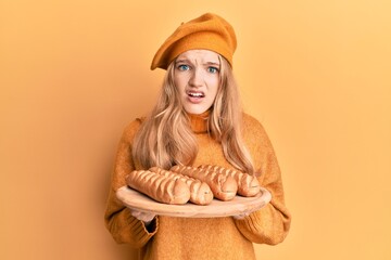 Beautiful young caucasian girl wearing french look with beret holding baguettes bread in shock face, looking skeptical and sarcastic, surprised with open mouth