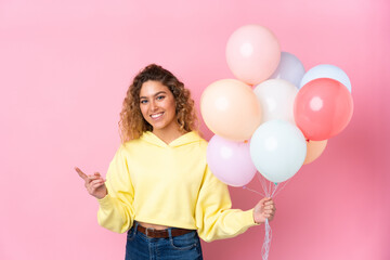 Young blonde woman with curly hair catching many balloons isolated on pink background pointing finger to the side