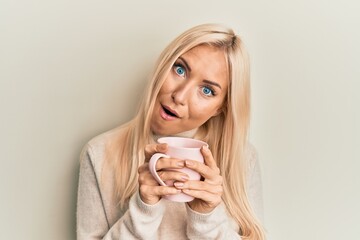 Young blonde woman drinking a cup of coffee in shock face, looking skeptical and sarcastic,...