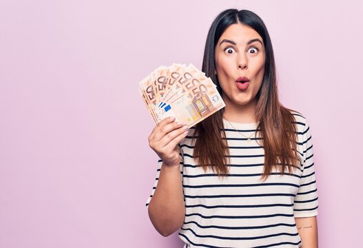 Young beautiful brunette woman holding euros banknotes over isolated pink background scared and amazed with open mouth for surprise, disbelief face
