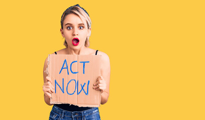 Young beautiful blonde woman holding act now banner scared and amazed with open mouth for surprise, disbelief face