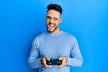 Young arab man playing video game holding controller winking looking at the camera with sexy expression, cheerful and happy face.