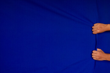 pair of a man's hands grasping a curtain and clenching his fists, as if he were lifting the blue fabric or dragging it, blue background with plenty of room for text