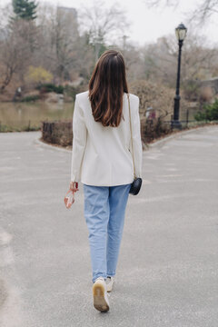 Stylish Young Woman Casual Suit Fashion Look. Wearing White Blazer, Light Blue Jeans, Sneakers. Back View, Walking In The Park, Blog Concept