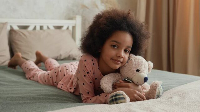 Cute little African American girl hugs her favorite teddy bear. Teenage girl in pajamas lies on bed in bedroom with beautiful light interior. Slow motion. Close up.