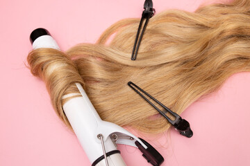 Obraz premium Curling blonde hair on a large diameter curling iron on a pink background. Curl care, hair styling, black clips.