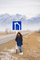 Travel destination. Caucasian girl with dreadlocks leans on blue road sign on snowy mountains background