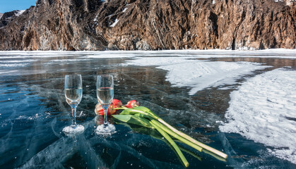 On clear, transparent blue ice, there are two champagne glasses and three red and yellow tulips. Visible texture of ice, cracks. Nearby there are areas of snow. The background is rocks. Lake Baikal