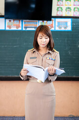 female thai teacher in government uniform standing in front of classroom reading an open note book
