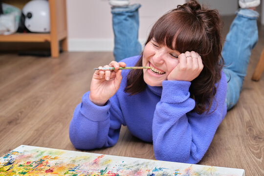 Young woman laughing, biting her paintbrush and painting on canvas with oil paints at home. Artist drawing laying on the floor.