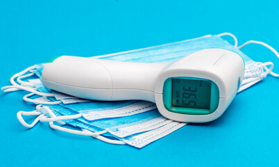 Electronic infrared thermometer with medical masks on blue background.