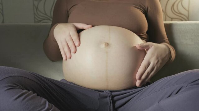 young pregnant woman stroking big belly on couch, making heart shape with hands