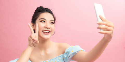 Image of beautiful brunette woman laughing and showing peace sign while taking selfie photo on cellphone isolated over pink background