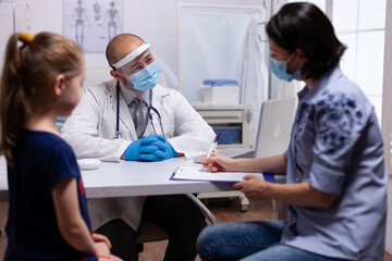 Fototapeta premium Mother completing medical questionnaire at doctor office in hospital clinic. Pediatrician with protection mask and visor against covid19 talking about diagnostic resultes providing care services