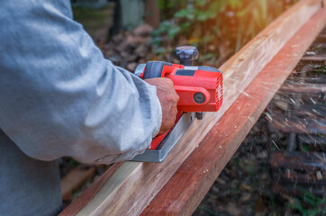 Hand of carpenter using electric planer with wooden plank in carpentry workshop in Thailand.