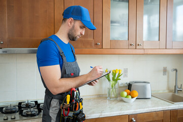 The repairman stands in the kitchen in a blue cap and writes in a tablet