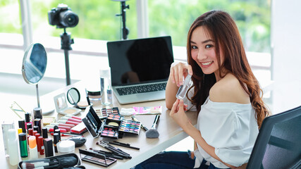 Young and cute Asian female vlogger, influencer, or online seller sitting with cosmetics products and DSLR camera and laptop notebook computer ready to broadcast online live stream or recording video