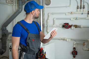 Young worker in a protective suit uses a tablet while checking the boiler equipment in the house.
