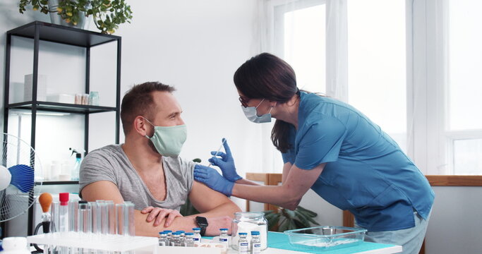 Vaccination Process. Young Nurse Doctor Woman In Blue Scrubs Gives Vaccine Injection Shot To Male Patient At Clinic.