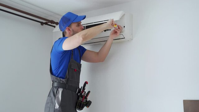 Male technician in overalls and a blue cap repairs an air conditioner on the wall