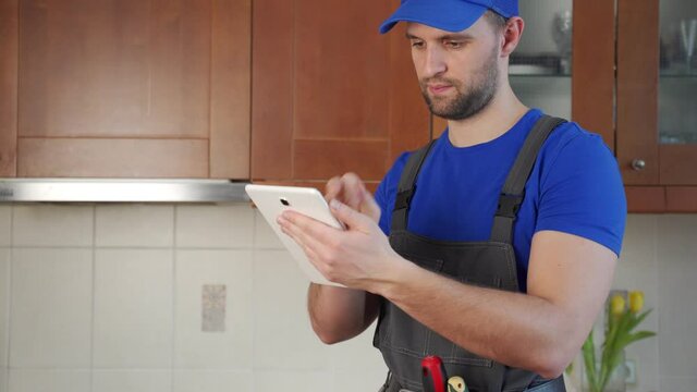 Plumber wearing a tool belt with various tools using tablet during work in kitchen