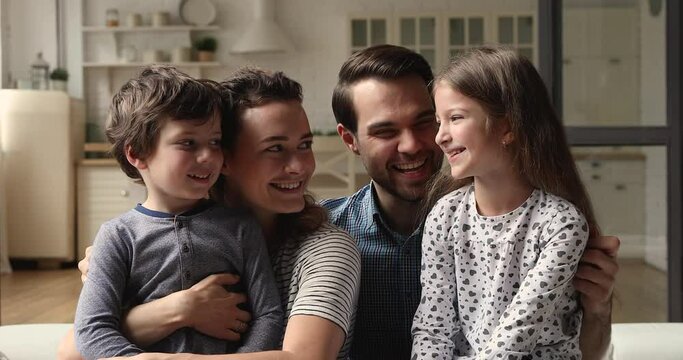 Little children sit on parents laps smiling look at camera, happy couple having fun play with cute daughter and son tickling them laughing enjoy time together. Beautiful young family portrait concept