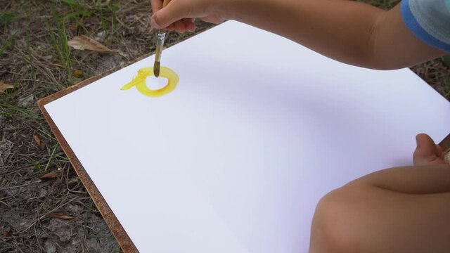 Closeup view video footage of cute drawing of little 5 year old baby. White preschool boy learning to draw sun using brush and yellow paints. White paper laying on ground and kids hand holding brush