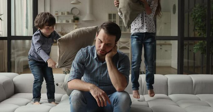 Tired man feels annoyed and fatigued due little daughter and son jumping on couch beating dad with cushions want play game. Hyperactive disobedient kids, difficulties of upbringing, quarantine concept