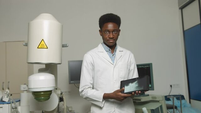 Doctor holding digital tablet pc. Close up portrait of smiling handsome African man doctor, standing in front of ultrasound and lithotripter machines. Safety non-invasive stone treatment.