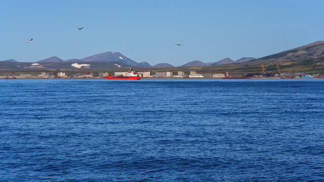 Summer seascape. A cargo ship near the coast. View of an abandoned northern settlement and an operating cargo seaport. In the distance tundra and mountains. Bering Sea. Beringovsky, Chukotka, Russia.
