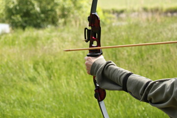 close-up of a hand holding a bow and aiming at a target, in the background a green landscape, a boy...