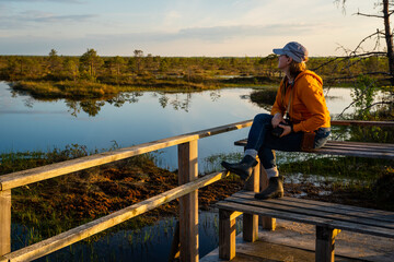 A female photographer admires the landscape of a forest lake among the raised bogs at sunset.