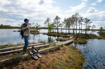 A man admires the landscape of a forest lake among the raised bogs at sunset. Nearby are devices for moving through the swamp. 