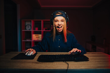 Portrait of a joyful lady gamer in casual clothes playing games at home on the computer at night and hiding from joy in a headset with a happy face. Positive female gamer playing.