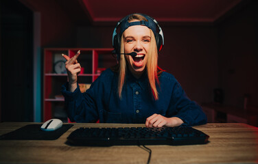 Happy female streamer in headset playing video games at home on computer looks at camera with happy...