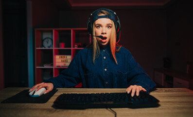 Fototapeta na wymiar Portrait of shocked female streamer in headset playing games at home on computer and looking at camera with shocked face.