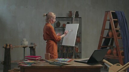 Professional female artist teaching art lesson online using laptop pc, live streaming tutorial video for vlog, explaining artistic techniques and skill of oil painting while working in art studio.