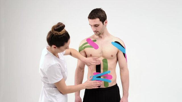 Female therapist applying kinesiology tape on a man's abdomen on the light background. Woman prepares male patient to glue kinesio adhesive tape on his belly. 4K