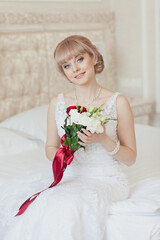 Pretty young Bride in elegant wedding dress with her bouquet. Blonde-haired woman with wedding hair-style in royal room of hotel. Boudoir morning of the bride in luxury interior. Wedding day ceremony