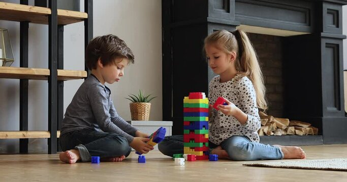 Siblings sit on warm floor in cozy living room with fireplace play plastic cubes. Little girl and boy enjoy playtime together build use colourful constructor bricks. Weekend leisure activity concept