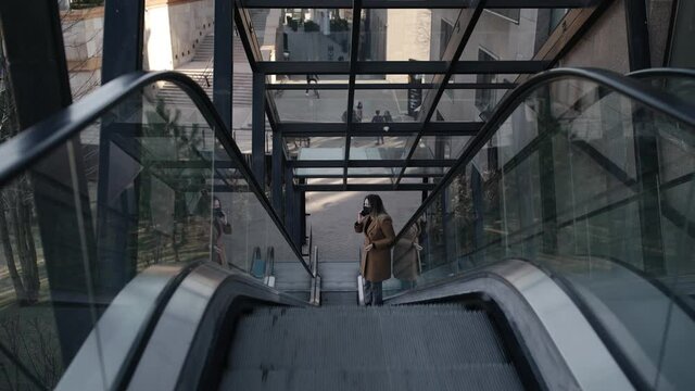 young blonde woman in a black mask dressed in a beige coat walking on a moving staircase or escalator speaking on the phone