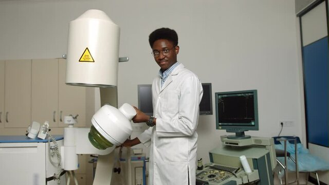 Portait of handsome young confident focused African man doctor, wearing white uniform, working in modern clinic with ultrasonic lithotripter machine. Kidney stones treatment concept