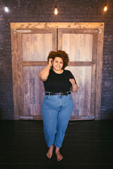 Beautiful curvy oversize African black woman afro hair barefoot in black t-shirt and jeans in loft interior with wooden door. Body imperfection, body acceptance, body positive and diversity concept