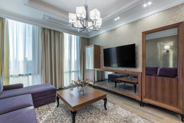 Spacious and fashionable light living room with comfortable violet sofa, coffee table and big french windows. A TV above the wooden cabinet and a wooden wardrobe.