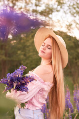 Young attractive blonde in a straw hat with a bouquet of lupins, among a purple field. The concept of nature and romance.